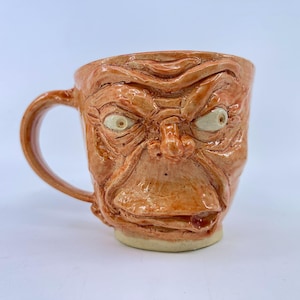 May include: A ceramic mug with a glossy orange finish, featuring a sculpted, wrinkled face with prominent eyes, a nose, and a wide mouth. The mug has a curved handle on the left side and a smooth, light-colored base, showcasing detailed textures.