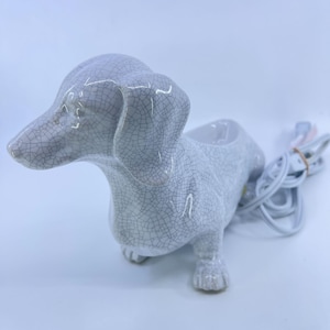 May include: A ceramic dachshund-shaped lamp featuring a white glaze with a dark gray crackle pattern. The dog figure has a hollowed-out back section and includes a white electrical cord with a plug extending from the rear of the body.