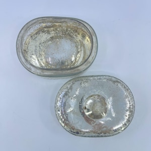 May include: An oval-shaped, clear glass container with a matching lid. The glass has a distressed, aged appearance with silver-colored accents. The lid features a small, round knob for easy handling. The container is likely for decorative or storage purposes.
