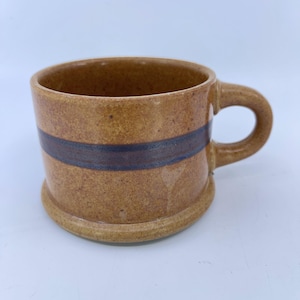 May include: A white ceramic mug with a black handle and the text "But First, Coffee" in a bold, black font. The mug is set against a blurred background, highlighting its clean design.