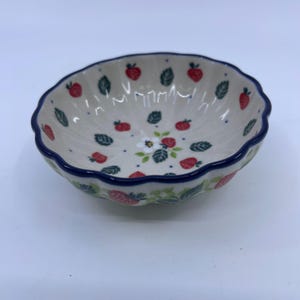 May include: A small, round, scalloped-edge ceramic bowl with a cream-colored base. The interior features a repeating pattern of small red strawberries, green leaves, and tiny blue dots, with a central white flower and green foliage design.