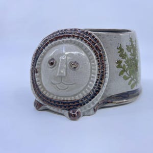 May include: A ceramic planter shaped like a lion with a light gray speckled finish. The lion face features a dark brown textured mane and paws. The side of the planter displays a green botanical illustration of a leafy plant with small white flowers.