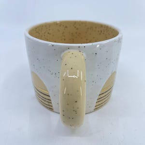 May include: White ceramic mug with a speckled exterior and a beige interior. The mug features a beige handle and decorative gold and black accents. The mug is designed for hot beverages.