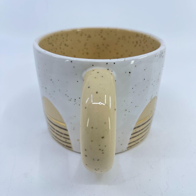 May include: White ceramic mug with a speckled exterior and a beige interior. The mug features a beige handle and decorative gold and black accents. The mug is designed for hot beverages.