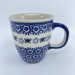 May include: A ceramic mug with a blue floral pattern on a cream background. The mug has a rounded shape with a blue handle and rim. The design includes stylized flowers and geometric shapes.
