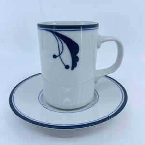 May include: White ceramic mug and saucer set. The mug has a dark blue rim and a decorative blue floral design. The saucer has a matching blue border. The set is designed for serving hot beverages.