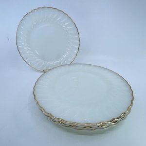 May include: Two white plates with a gold trim. The plates have a spiral design and a scalloped edge. The plates are stacked on top of each other. The plates are made of a shiny material.