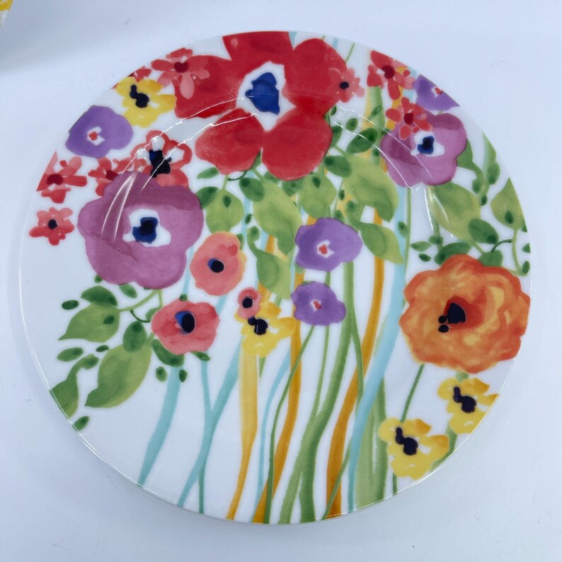 May include: A white ceramic plate with a colorful floral design. The plate features red, purple, orange, and yellow flowers with green leaves and blue and yellow stems. The flowers are painted in a watercolor style.