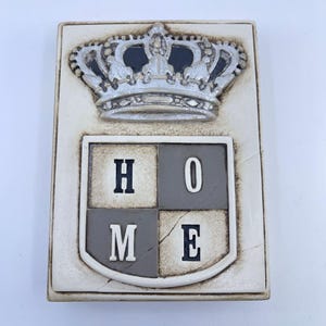 May include: A rectangular, off-white plaque with a silver crown at the top and the word "HOME" in black letters on a shield below. The crown is detailed with black accents, and the shield is divided into four sections.