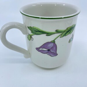 Puede incluir: Taza de cer&aacute;mica blanca con borde y asa verdes. La taza presenta un dise&ntilde;o de flor morada y hojas verdes. Tambi&eacute;n se ve una peque&ntilde;a mariposa.