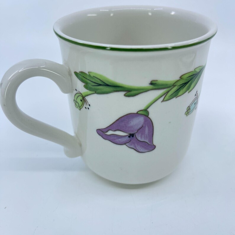Puede incluir: Taza de cer&aacute;mica blanca con borde y asa verdes. La taza presenta un dise&ntilde;o de flor morada y hojas verdes. Tambi&eacute;n se ve una peque&ntilde;a mariposa.