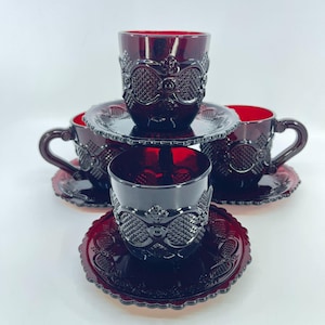 May include: A set of four deep red glass cups and saucers featuring a raised diamond and scroll pattern. The cups have curved handles and the saucers have scalloped edges. The glassware has a translucent finish and a pressed texture throughout.
