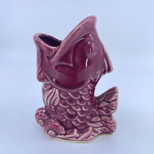 May include: A maroon ceramic fish-shaped vase with an open mouth. The vase has a textured, scaled body and fins. The interior of the mouth is dark, and the vase sits on a light-colored base.