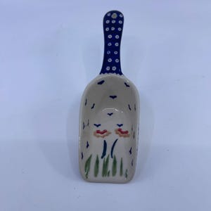 May include: A ceramic scoop featuring a cream-colored bowl decorated with hand-painted green grass, orange flowers, and small blue bird motifs. The handle is dark blue with a repeating pattern of white dots and a small hole at the very top end.