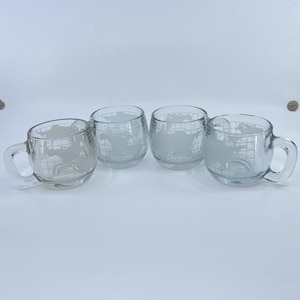 May include: Four clear glass mugs featuring a frosted white etched design of a world map with longitudinal and latitudinal grid lines. Each mug has a rounded bowl shape and a sturdy handle, displaying a continuous global map pattern.