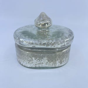 May include: A small, silver-colored jar with a textured surface and a matching lid. The lid is topped with a small, pointed finial. The jar is likely made of glass or a similar material, and it has a vintage or antique appearance.