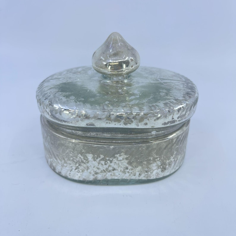 May include: A small, silver-colored jar with a textured surface and a matching lid. The lid is topped with a small, pointed finial. The jar is likely made of glass or a similar material, and it has a vintage or antique appearance.