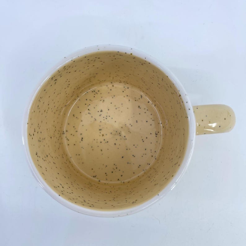 May include: A speckled ceramic mug with a cream-colored interior and handle. The interior features small, dark brown speckles. The exterior rim is white. The mug is viewed from above, showcasing the inside and handle.
