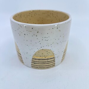 May include: A white ceramic cup with a speckled exterior and a tan interior. The cup features a decorative design of tan arches with gold stripes and brown horizontal lines. The cup is a unique piece of pottery.