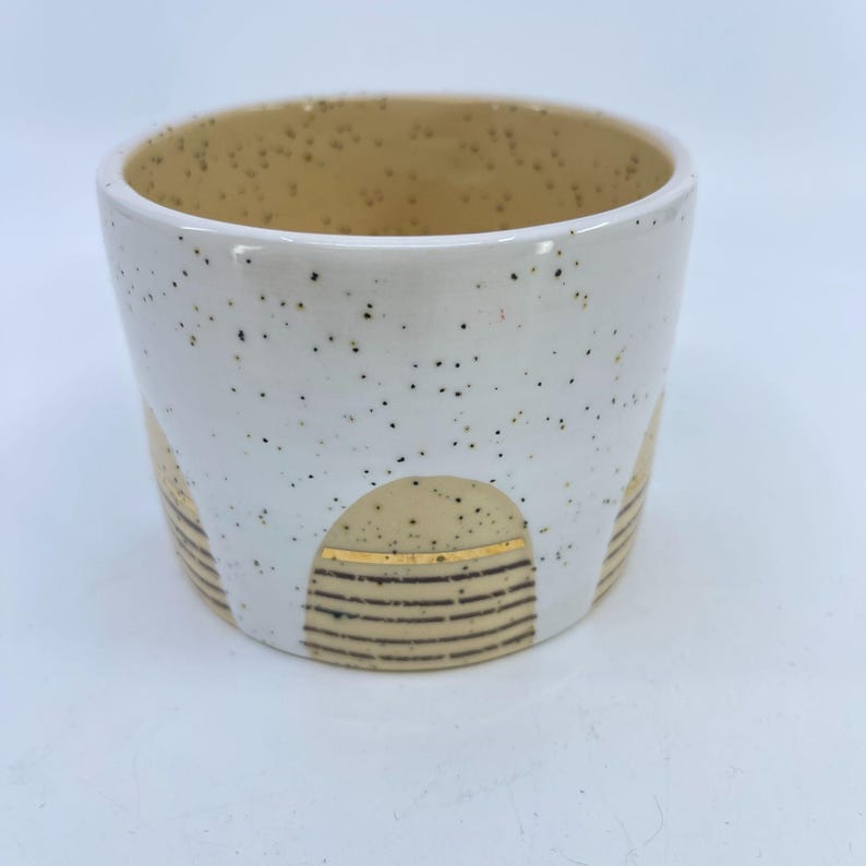 May include: A white ceramic cup with a speckled exterior and a tan interior. The cup features a decorative design of tan arches with gold stripes and brown horizontal lines. The cup is a unique piece of pottery.