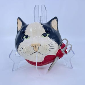 May include: A ceramic cat head ornament featuring black and white markings, green eyes, and a red scarf around the neck. A pair of silver-toned metal scissors is tucked into the scarf, and a thin white string hangs from the cat's nose area.