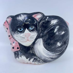 May include: A ceramic cat-shaped dish with a black and white coat, blue eyes, and a pink nose. The cat is curled up, and a pink scarf with black polka dots is around its neck. The dish is likely for decorative or functional use.