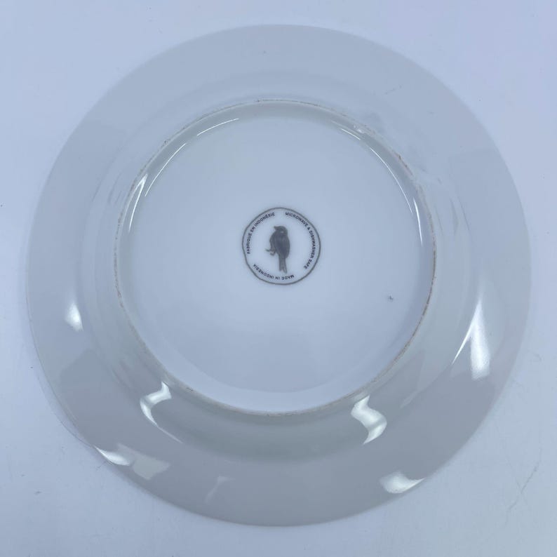 May include: White ceramic plate with a slightly raised rim. The center of the plate features a small, circular design with a bird silhouette and text. The plate is designed in Indonesia.