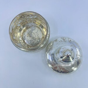 May include: A clear glass jar with a lid, both featuring a distressed, silver mercury glass finish. The jar is round with a wide opening, and the lid has a knob. The jar and lid are separated, showing the interior.