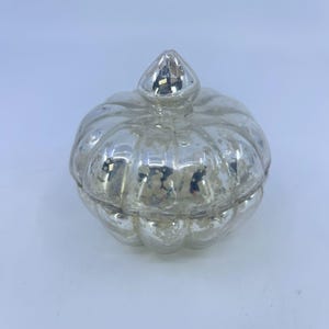 May include: A silver-coloured, pumpkin-shaped decorative jar with a lid. The jar has a textured, distressed finish, and the lid is topped with a small, pointed finial. The jar is likely made of glass or a similar material.