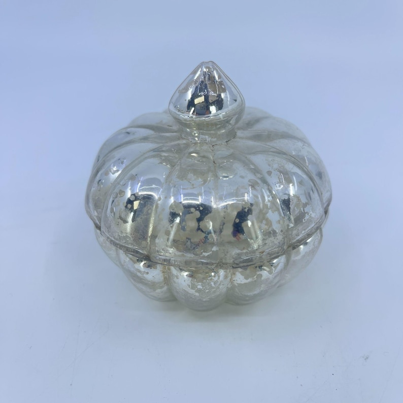May include: A silver-coloured, pumpkin-shaped decorative jar with a lid. The jar has a textured, distressed finish, and the lid is topped with a small, pointed finial. The jar is likely made of glass or a similar material.
