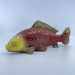 May include: A ceramic fish sculpture with a maroon body, yellow head, and green fins. The fish has a textured, scaled pattern and is displayed on a white surface. This decorative piece could be used as a home decor accent.