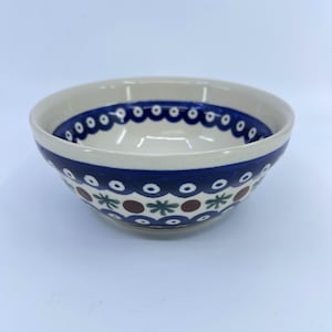 May include: A ceramic bowl with a cream-colored interior and a blue and white patterned exterior. The rim features a dark blue scalloped design, while the body displays alternating patterns of brown dots, green leaves, and blue circles.