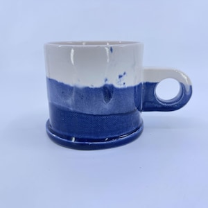 May include: A ceramic mug featuring a two-tone design with a white upper section and a deep blue lower section. The mug has a cylindrical body with a textured, dimpled surface and a thick, circular handle extending from the right side.