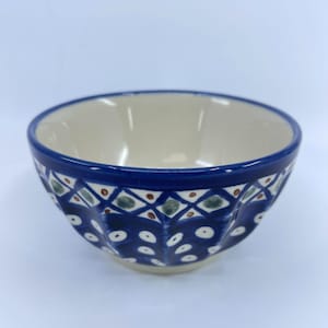 May include: A ceramic bowl with a cream-colored interior and a dark blue exterior rim. The bowl's exterior features a repeating pattern of white, green, and brown geometric shapes. The bowl is likely used for serving food.