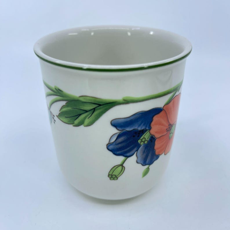 Puede incluir: Una taza de cer&aacute;mica blanca con borde verde y dise&ntilde;o floral. Las flores son en tonos azules y coral, con hojas y tallos verdes. La taza tiene una forma suave y redondeada.