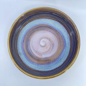 May include: A handmade ceramic bowl with a spiral design. The bowl features a color palette of brown, blue, and purple hues. The rim is a warm brown, transitioning to blue and purple in the center, creating a unique, artistic piece.