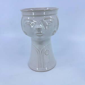 May include: A glossy off-white ceramic vessel shaped like a stylized human head and torso. The piece features embossed facial features, a wide circular rim, and vertical line detailing on the neck area, creating a smooth, uniform finish.