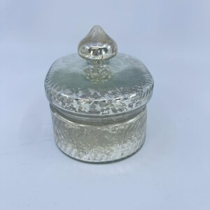 May include: A small, round, silver-colored jar with a lid. The jar and lid have a textured, hammered appearance. The lid is topped with a small, teardrop-shaped knob. The jar is likely made of glass or a similar material.