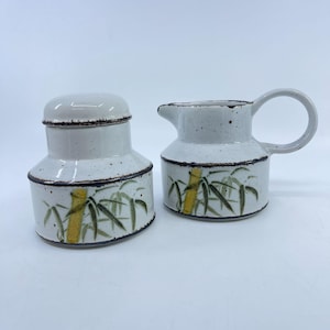 May include: A ceramic sugar bowl with a lid and a matching creamer pitcher. Both pieces are off-white with a speckled finish, featuring a dark brown rim and a hand-painted design of green bamboo leaves and yellow stalks around the base.