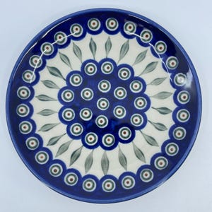 May include: A round ceramic plate with a blue rim and a central floral design. The plate features a white background with a pattern of blue circles, green leaves, and red and green dots. The plate is a decorative piece.