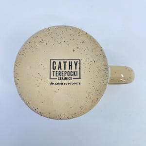 May include: A ceramic mug with a speckled beige exterior and a curved handle. The bottom of the mug features a rectangular black and white label with the text "CATHY TEREPOCKI CERAMICS for ANTHROPOLOGIE".