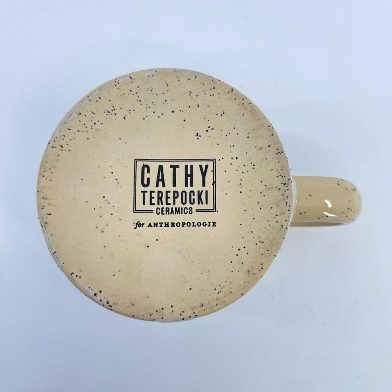 May include: A ceramic mug with a speckled beige exterior and a curved handle. The bottom of the mug features a rectangular black and white label with the text "CATHY TEREPOCKI CERAMICS for ANTHROPOLOGIE".