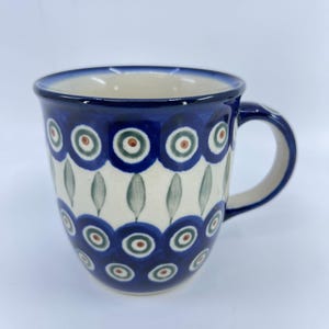 May include: A ceramic mug with a blue handle and rim. The mug features a white base with a repeating pattern of blue circles, green and brown accents, and stylized green leaves. The mug is designed for hot beverages.