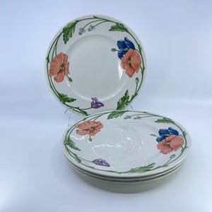 May include: White ceramic plates with a floral design. The plates feature orange, blue, and purple flowers with green leaves around the rim. The plates are stacked and displayed on a white background.