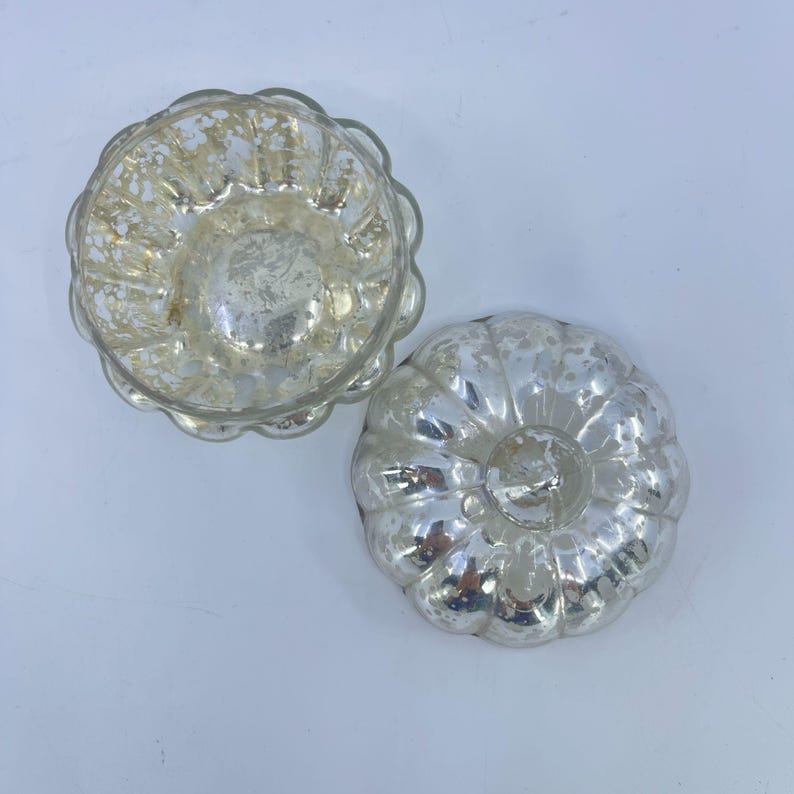 May include: Two silver-coloured, vintage-style glass moulds. One is open, revealing the interior, and the other is upside down. The moulds have a fluted, pumpkin-like shape and a distressed, antique finish. They are likely used for baking or decorative purposes.