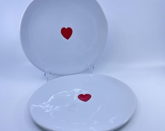 May include: Two round white ceramic plates, each featuring a single small red heart centered on the surface. One plate is positioned upright on a clear stand, while the second plate lies flat in front of it, both displaying a smooth finish.