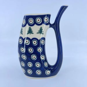 May include: A ceramic horn-shaped drinking vessel featuring a deep blue base color with a white band near the top. The surface is decorated with repeating dark blue and white circular patterns and small green pine tree motifs throughout.