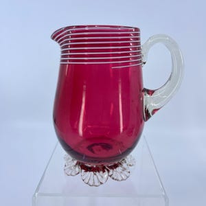 May include: A ruby red glass pitcher with a clear handle and a decorative clear base. The pitcher has horizontal white stripes around the top. The pitcher is likely used for serving beverages.