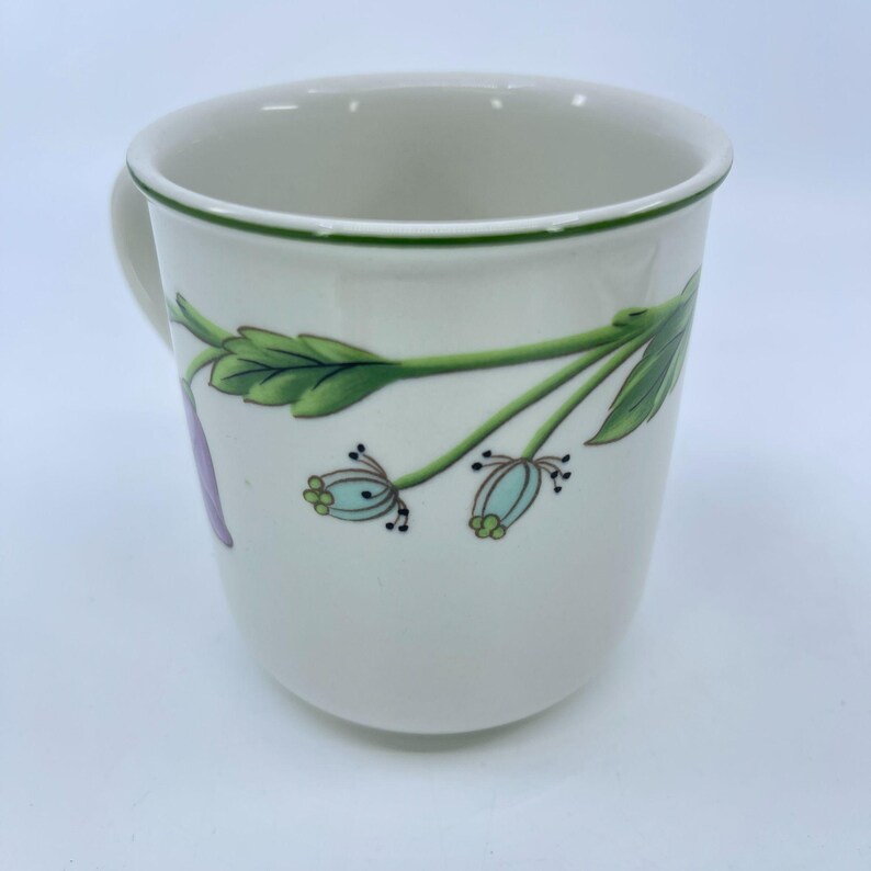 Puede incluir: Taza de cer&aacute;mica blanca con borde y asa verdes. La taza presenta un dise&ntilde;o floral con hojas verdes, capullos de flores azul claro y un p&eacute;talo morado. La taza es de tama&ntilde;o est&aacute;ndar para caf&eacute; o t&eacute;.