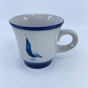 May include: White ceramic mug with a blue sailboat design. The mug has a light gray interior and a dark blue rim and base. The handle is light gray.